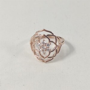 May include: A rose gold ring featuring an openwork lotus flower design. The centre of the floral motif is embellished with a cluster of six small, clear, round-cut stones arranged in a hexagonal pattern, set within the metallic frame.