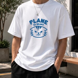 May include: A white short-sleeved t-shirt featuring a blue graphic print on the chest. The graphic displays the text "PLANE IS READY" in a bold blue font above a stylized, grumpy-looking cat face illustration in blue ink.