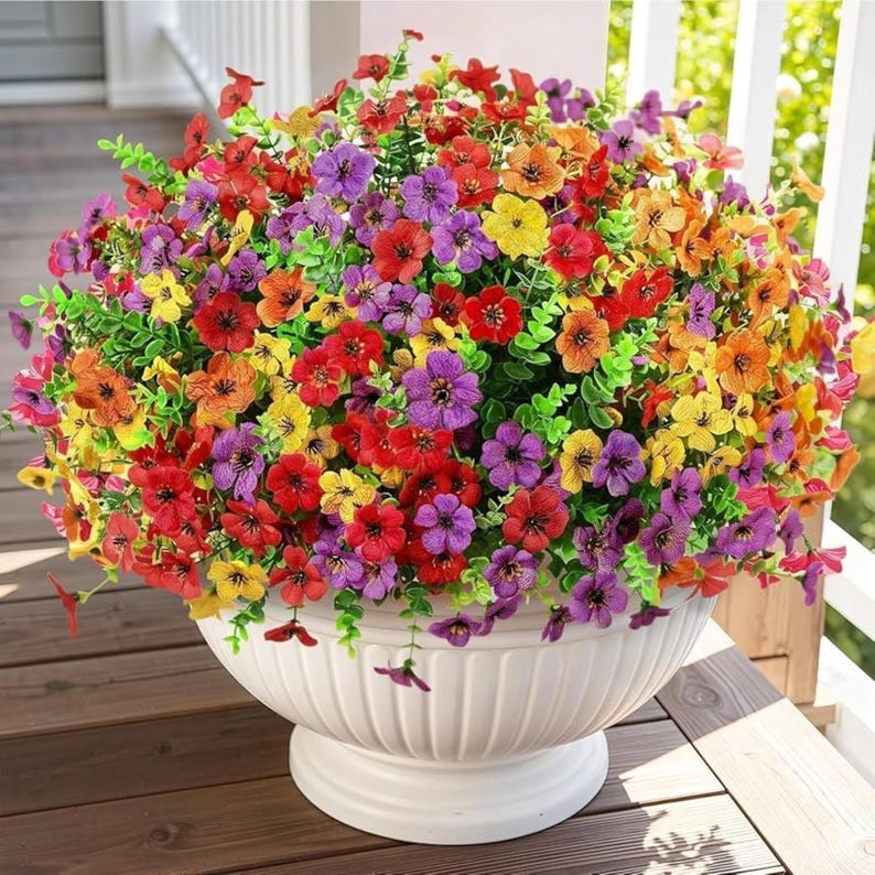 K&ouml;nnte beinhalten: Ein gro&szlig;es, rundes Arrangement aus k&uuml;nstlichen Blumen in leuchtenden Rot-, Orange-, Gelb- und Violettt&ouml;nen mit kleinen f&uuml;nfbl&auml;ttrigen Bl&uuml;ten und gr&uuml;nem Blattwerk, pr&auml;sentiert in einer wei&szlig;en, geriffelten Schale auf einem Fu&szlig;.