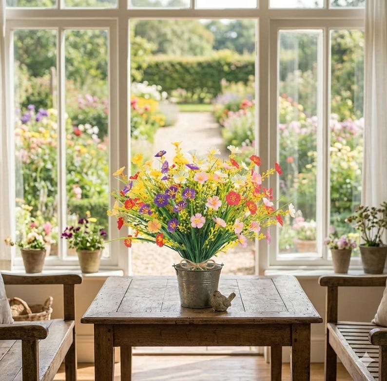 Pode incluir: Um balde de metal preenchido com um arranjo vibrante de flores silvestres artificiais em tons de amarelo, roxo, rosa, laranja e vermelho. O balde repousa sobre uma mesa de madeira, com caules e folhagens verdes que criam um arranjo denso.
