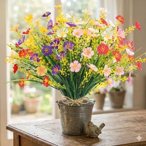 Pode incluir: Um ramo de flores silvestres artificiais em tons de amarelo, roxo, rosa e vermelho, com caules e folhagem verdes. As flores est&atilde;o dispostas num pequeno balde de metal cinzento desgastado com um la&ccedil;o de juta e uma pequena figura de p&aacute;ssaro.