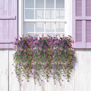 May include: A window box overflowing with artificial trailing flowers in shades of purple, violet, and pink, featuring small five-petaled blossoms and green foliage, mounted beneath a white window frame with light purple wooden shutters.