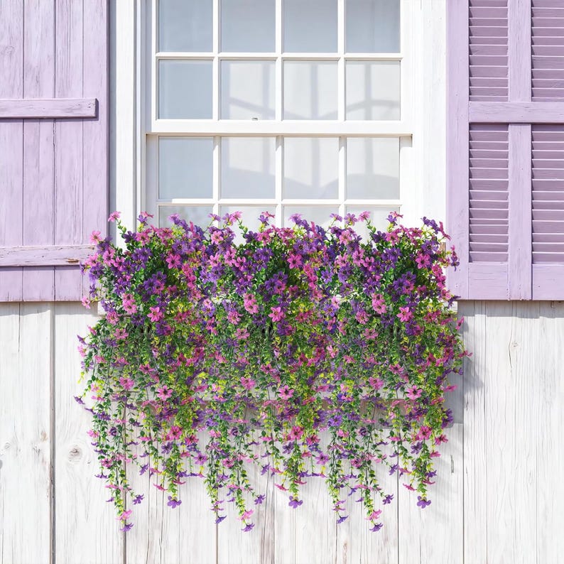 May include: A window box overflowing with artificial trailing flowers in shades of purple, violet, and pink, featuring small five-petaled blossoms and green foliage, mounted beneath a white window frame with light purple wooden shutters.
