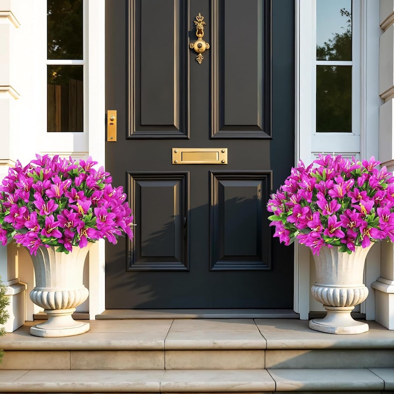 May include: Two off-white pedestal planters containing vibrant magenta flowers with green leaves, positioned on either side of a dark charcoal gray front door featuring a gold-colored metal door knocker and a rectangular mail slot.
