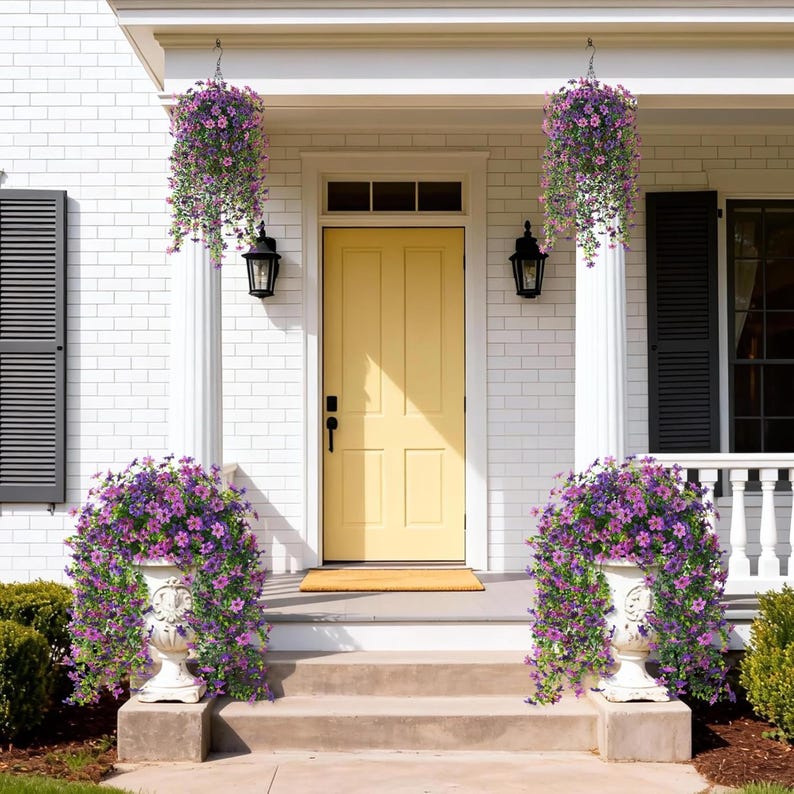 May include: A front porch featuring a yellow door, two hanging baskets filled with purple flowers, and two white pedestal planters overflowing with matching purple flowers. The house has white brick siding, black shutters, and two black wall lanterns.