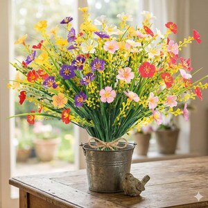 Pode incluir: Um ramo de flores silvestres artificiais em tons de amarelo, roxo, rosa e vermelho, com caules e folhagem verdes. As flores est&atilde;o dispostas num pequeno balde de metal cinzento desgastado com um la&ccedil;o de juta e uma pequena figura de p&aacute;ssaro.