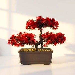 Op de afbeelding: Een decoratieve kunstmatige bonsaiboom met levendig rood blad en een donkerbruine, getextureerde stam. De boom is geplant in een rechthoekige, donkerbruine keramische pot die aan de basis is gevuld met kleine, lichtgekleurde steentjes.