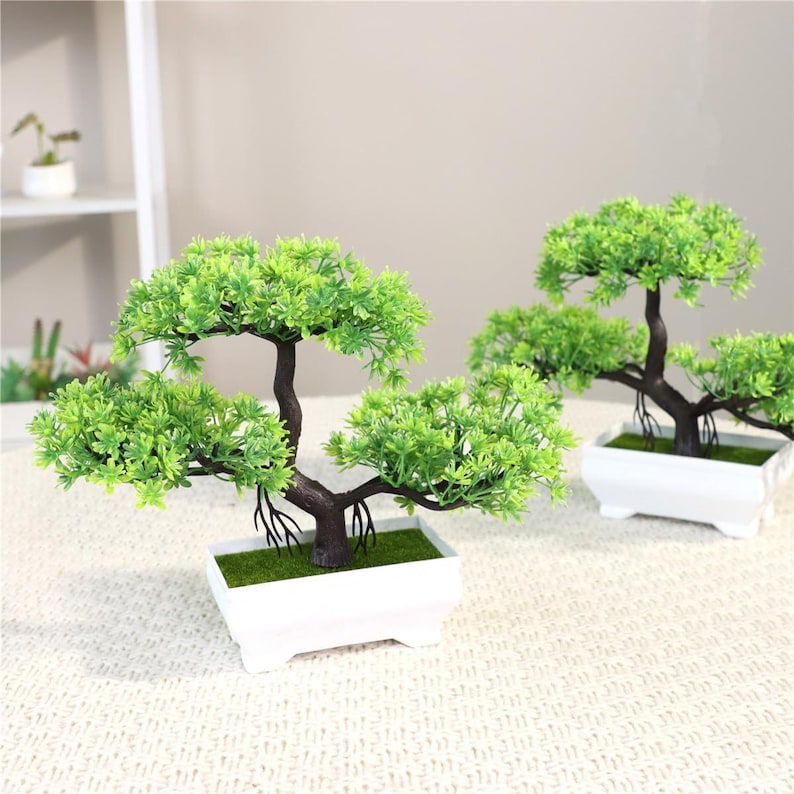 May include: Two artificial bonsai trees featuring vibrant green plastic foliage and dark brown textured trunks. Each tree is potted in a rectangular white plastic container filled with a layer of green faux moss at the base of the trunk.