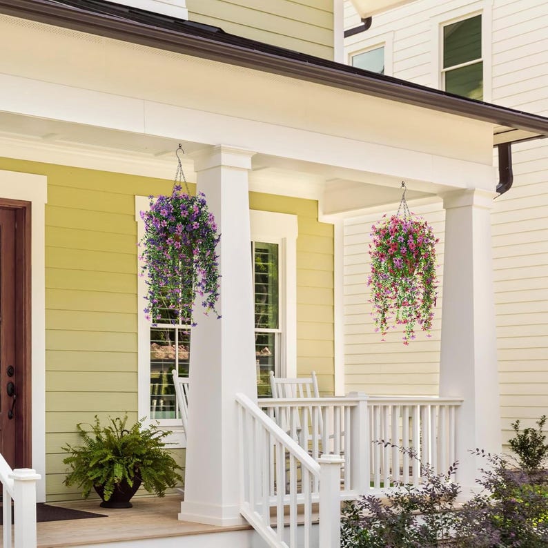 May include: Two hanging baskets filled with cascading purple and pink flowers suspended from the ceiling of a porch. Each basket features lush green foliage and vibrant blooms, creating a decorative display on the front of a house.