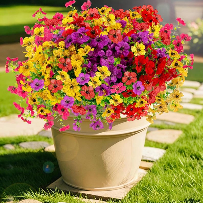 K&ouml;nnte beinhalten: Ein gro&szlig;er, runder, beigefarbener Kunststoff-Pflanzk&uuml;bel, gef&uuml;llt mit einem dichten Arrangement aus k&uuml;nstlichen Blumen in leuchtenden Rot-, Gelb- und Violettt&ouml;nen. Die Blumen haben jeweils f&uuml;nf Bl&uuml;tenbl&auml;tter und sind mit kleinen gr&uuml;nen Stielen und Bl&auml;ttern durchsetzt.