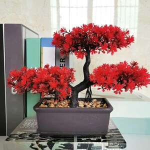 Op de afbeelding: Een decoratieve kunstmatige bonsaiboom met levendig rood blad en een donkerbruine, getextureerde stam. De boom is geplant in een rechthoekige, donkerbruine pot gevuld met kleine, lichtgekleurde steentjes voor een gestructureerde en gebalanceerde uitstraling.