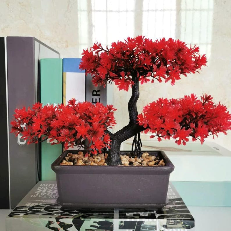 Op de afbeelding: Een decoratieve kunstmatige bonsaiboom met levendig rood blad en een donkerbruine, getextureerde stam. De boom is geplant in een rechthoekige, donkerbruine pot gevuld met kleine, lichtgekleurde steentjes voor een gestructureerde en gebalanceerde uitstraling.