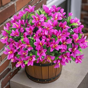 May include: A dense arrangement of vibrant magenta flowers with yellow centers and green leaves, housed in a round, brown wooden barrel-style planter featuring two horizontal black metal bands with small rivets around the exterior surface.