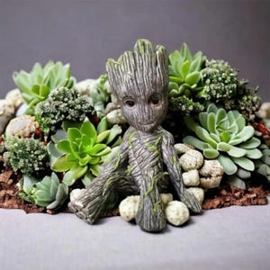 May include: A small, grey, tree-like figurine with a textured bark surface and subtle green moss accents, seated among a cluster of various green succulent plants and small, light-colored, rounded stones on a flat, neutral-toned surface.