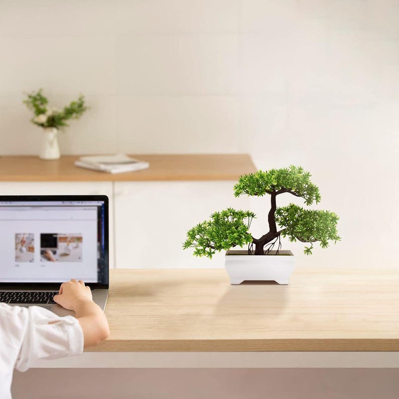 May include: A small artificial bonsai tree with vibrant green foliage and a dark brown trunk, potted in a rectangular white ceramic container. The plant features multiple layered branches and is positioned on a light-colored wooden desk surface.