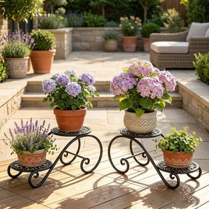 May include: Two black metal plant stands with scrollwork legs, each holding two potted plants. The left stand holds lavender and purple hydrangeas in terracotta pots. The right stand holds pink hydrangeas and green foliage in patterned pots.