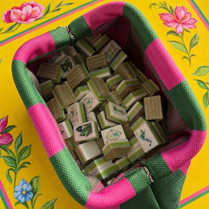 Könnte beinhalten: Ein Stoffbeutel mit einem Karomuster aus abwechselnden rosa und grünen Quadraten, gefüllt mit einer Sammlung rechteckiger Mahjong-Spielsteine. Die Spielsteine haben hellgrüne und weiße Oberflächen mit verschiedenen Symbolen und Abbildungen.