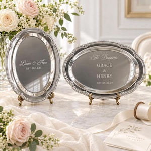 Puede incluir: Dos bandejas ovaladas de tono plateado con bordes de cuentas ornamentados, cada una apoyada sobre pequeños soportes de metal. La bandeja vertical está grabada con "Liam & Ava" y "EST 08.14.27". La horizontal tiene "The Bennetts", "GRACE & HENRY" y "EST 05.30.27".