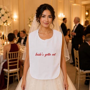 Può includere: Una bavaglino in tessuto bianco con il testo ricamato in rosso bride's gotta eat, indossato sopra un abito da sposa bianco con un velo trasparente. Il bavaglino presenta uno scollo arrotondato e un design semplice che copre la parte anteriore.