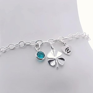 Puede incluir: Una pulsera de tobillo plateada con un diseño de eslabones. La pulsera de tobillo presenta tres dijes: una gema redonda de color verde azulado, un trébol de cuatro hojas y un corazón con la letra "E". Los dijes cuelgan de la cadena.