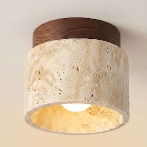 Könnte beinhalten: Eine zylindrische Deckenleuchte mit einer Basis aus dunkelbraunem Holz und einem Schirm aus strukturiertem, porösem beigem Travertinstein. Die Lichtquelle ist im Inneren des Steinzylinders sichtbar und strahlt ein warmes Licht aus.