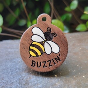 Puede incluir: Una etiqueta redonda de madera para mascotas con una ilustración de abeja negra y amarilla y el texto "Buzzin'" grabado en ella.