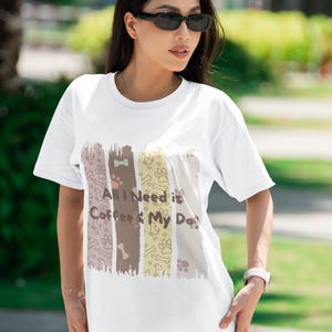 Puede incluir: Una camiseta blanca de manga corta con un diseño gráfico de cuatro paneles verticales de pinceladas en colores suaves. El diseño incluye huellas de patas, formas de huesos y el texto "All I Need is Coffee & My Dog" en letra marrón oscuro.