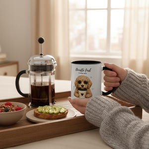 May include: A white ceramic mug with a black rim and handle, featuring a graphic of a golden doodle puppy holding a coffee cup and the text Doodle First Coffee Later. The mug is held by a person wearing a grey sweater, next to a French press.