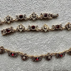 May include: A gold-toned metal chain necklace featuring a repeating pattern of circular deep red faceted stones and small white accents set within ornate, textured metal frames. The necklace includes a secure metal clasp closure at the end.