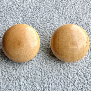 May include: Two circular wooden buttons with a smooth, polished finish. Each button features a light brown color with visible natural wood grain patterns running horizontally across the surface. The buttons are round, flat, and have a glossy appearance.