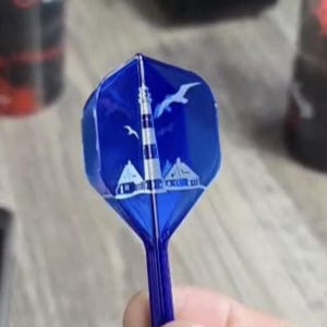 Könnte beinhalten: Ein blauer Dartpfeil-Flight mit einem weißen Leuchtturm-Design, zwei Möwen und zwei Häusern. Der Flight wird in einer Hand gehalten.