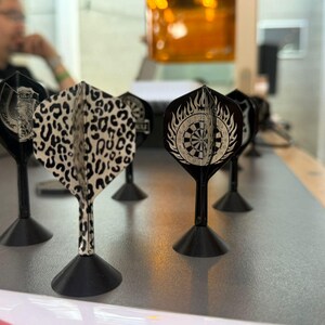 Könnte beinhalten: Eine Sammlung von Dartpfeil-Flights mit verschiedenen Designs, darunter ein Leopardenmuster und ein Ziel mit Flammen. Die Flights sind schwarz mit weißen und beigen Mustern. Sie sind auf schwarzen Schäften mit schwarzen Basen montiert, die auf einer grauen Oberfläche angeordnet sind.