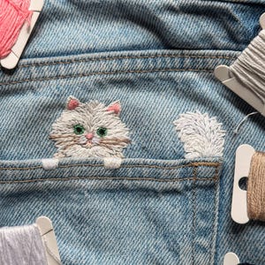 Könnte beinhalten: Eine hellblaue Jeanstasche mit einer Stickerei eines flauschigen Katzenkopfes mit grünen Augen und rosa Ohren, der über den Rand schaut, ergänzt durch einen weißen gestickten Schwanz, umgeben von mehreren Kunststoffspulen mit verschiedenfarbigem Stickgarn.