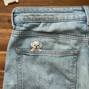 May include: Light blue denim jeans with a small, embroidered white dog peeking out of the back pocket. The dog has a fluffy appearance and is centered in the pocket. The jeans are slightly faded.