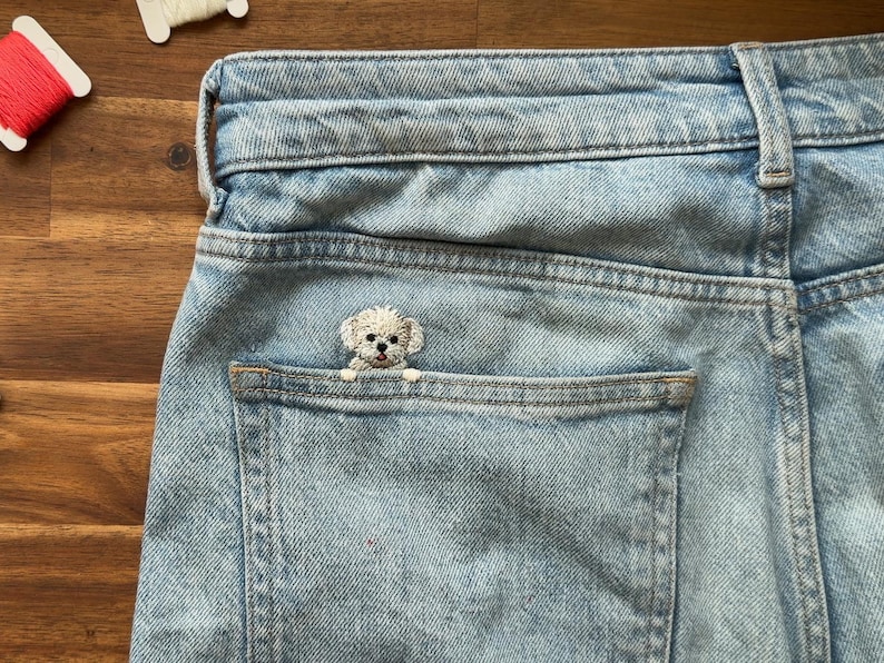 May include: Light blue denim jeans with a small, embroidered white dog peeking out of the back pocket. The dog has a fluffy appearance and is centered in the pocket. The jeans are slightly faded.