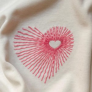 Könnte beinhalten: Ein cremefarbener Stoff mit einem gestickten rosa Herzmotiv. Die Stickerei besteht aus strahlenförmig angeordneten geraden Stichen, die eine große Herzform bilden, mit einem kleinen, nicht bestickten herzförmigen Freiraum in der Mitte.