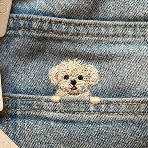 Könnte beinhalten: Nahaufnahme einer hellblauen Jeans-Tasche mit einem beigefarbenen, gestickten Welpen, der herauslugt. Der Welpe hat schwarze Augen und eine rote Zunge. Spulen mit weißem und rotem Faden sind sichtbar.