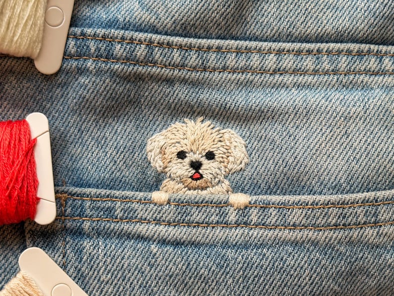 May include: Close-up of a light blue denim pocket with a beige embroidered puppy peeking out. The puppy has black eyes and a red tongue. Spools of white and red thread are visible.