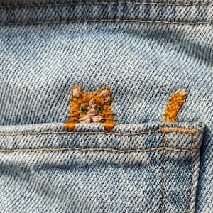 Può includere: Un tessuto in denim azzurro con il ricamo di un gatto tigrato arancione che spunta dal bordo superiore di una tasca. Il gatto ha baffi visibili, naso rosa e occhi verdi, con la coda anch'essa ricamata che appare sulla destra della testa.