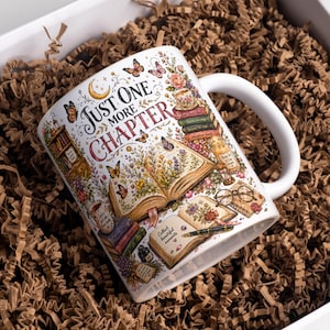May include: A white ceramic mug featuring a colourful illustration of open books, butterflies, flowers, and reading accessories. The text "JUST ONE MORE CHAPTER" is printed in a decorative font. Additional text includes "Collect beautiful moments" on a book page.