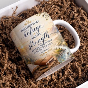 May include: A white ceramic mug featuring a printed landscape illustration of a river and mountains. The text reads: THE LORD IS MY refuge AND MY strength AN EVER-PRESENT HELP IN TIMES OF TROUBLE PSALM 46:1. A brown book and pen are depicted below.