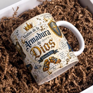 Könnte beinhalten: Ein weißer Keramikbecher mit einem detaillierten Grafikdesign, das den Text "Vestíos de la Armadura de Dios" und "Efesios 6:11" zeigt. Die Illustration umfasst eine goldene Krone, einen Schild, Sandalen und verschiedene religiöse Symbole in Gold, Blau und Schwarz.