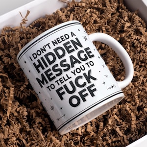 May include: A white ceramic mug featuring black text that reads, I DON'T NEED A HIDDEN MESSAGE TO TELL YOU TO FUCK OFF. The mug is decorated with small black geometric symbols and lines, including stars, arrows, and circles, arranged around the text.