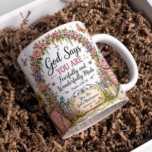 May include: A white ceramic mug featuring a colorful floral arch illustration with butterflies and birds. The text reads: God Says YOU ARE Fearfully and Wonderfully Made Psalm 139:14 and You Are God's Masterpiece Ephesians 2:10, with a lantern and garden gate.