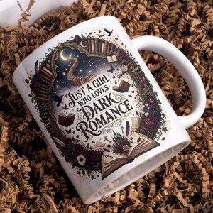 May include: A white ceramic mug featuring a detailed graphic print with the text "JUST A GIRL WHO LOVES DARK ROMANCE". The design includes an open book, a crescent moon, a staircase, a castle, dark roses, and flying birds in a circular frame.