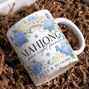 May include: A white ceramic mug featuring a floral design of blue and white hydrangeas, butterflies, and mahjong tiles. The text reads MAHJONG game night favorite and TILES & BLOOMS. The mug has a curved handle and is decorated with pearl strings.