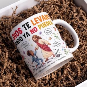 May include: A white ceramic mug featuring a colourful illustration of a figure in a robe helping a person stand up. The text reads DIOS TE LEVANTA CUANDO YA NO PUEDES, Yo Estoy Contigo, No Temas, Sigue Adelante, Ansiedad, Miedo, Duda, Culpa, Dolor, Preocupación.