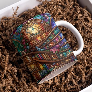 May include: A white ceramic mug featuring a colorful stained glass style illustration of curved bookshelves filled with books, lit candles, small owls, and floral accents. The design wraps around the mug, which has a plain white handle.