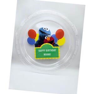 May include: A clear plastic plate featuring Sesame Street characters Cookie Monster and Elmo, with red, blue, and yellow balloons. A green sign reads "HAPPY BIRTHDAY MIANA!"