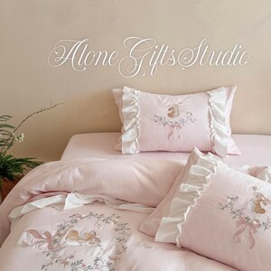 Può includere: Un set di biancheria da letto rosa chiaro composto da un copripiumino e due federe con bordi a volant bianchi. Il tessuto è ricamato con illustrazioni di conigli marroni, ghirlande floreali e fiocchi rosa. Il testo Alone Gifts Studio è visibile.